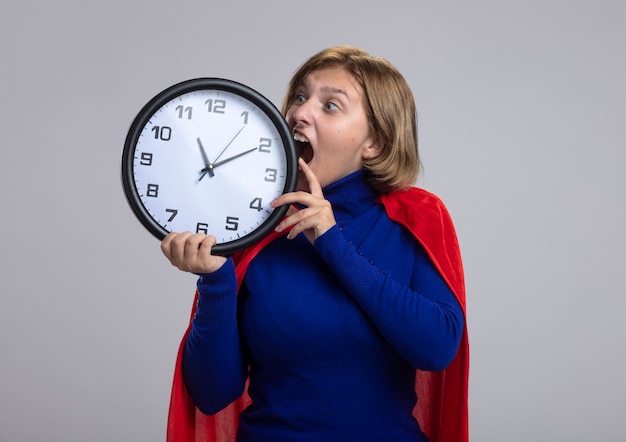 Молодая блондинка супергерой девушка в красном плаще держит и смотрит на часы, пытаясь укусить их, изолированные на белом фоне с копией пространства