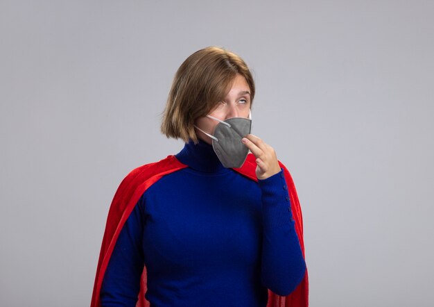 Young blonde superhero girl in red cape wearing protective mask trying to take it off rolling eyes isolated on white background with copy space