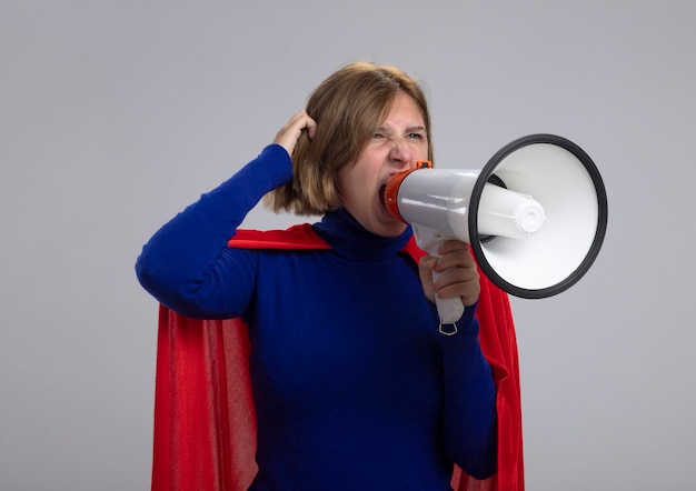 Young blonde superhero woman in red cape looking straight touching hair shouting in loud speaker isolated on white wall