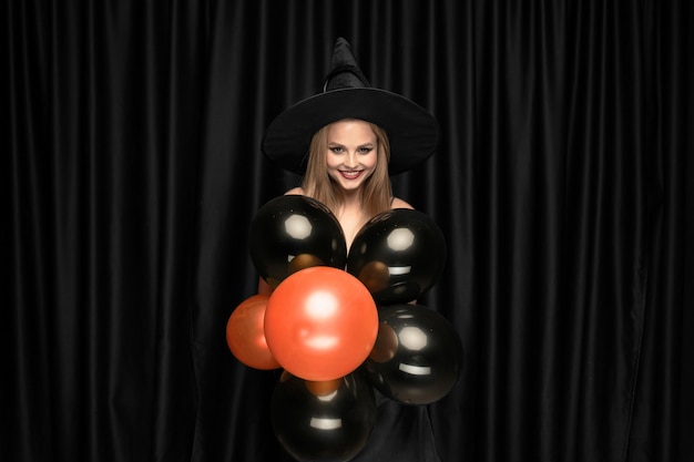 Young blonde woman in black hat and costume on black background. Attractive, sensual female model. Halloween, black friday, cyber monday, sales, autumn