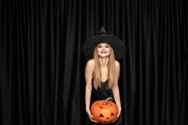 Young blonde woman in black hat and costume on black background. Attractive, sensual female model. Halloween, black friday, cyber monday, sales, autumn
