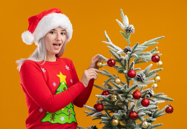 Молодая блондинка в рождественском свитере и шапке санта-клауса, украшающей елку, счастлива и позитивно улыбается, бодро улыбается елка