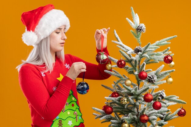 Young blonde woman in christmas  sweater and santa hat decorating christmas tree happy and positive smiling cheerfully 