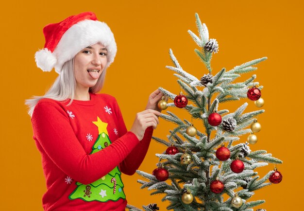 Young blonde woman in christmas  sweater and santa hat decorating christmas tree happy and positive sticking out tongue 