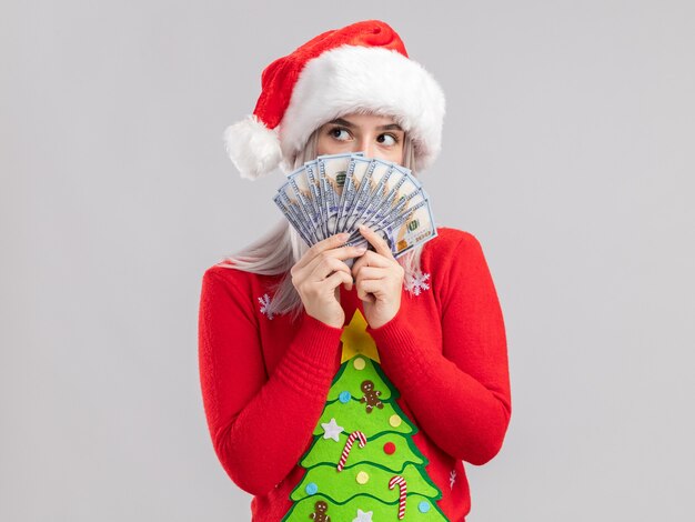 真面目な顔で脇を探して現金を保持しているクリスマスセーターとサンタ帽子の若いブロンドの女性