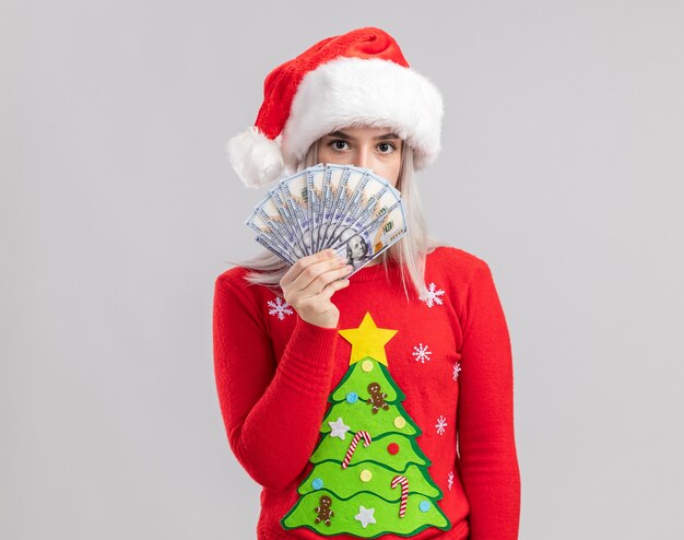 Foto gratuita giovane donna bionda in maglione di natale e cappello della santa che tiene contanti alla ricerca con la faccia seria