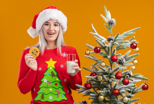 顔に笑顔で牛乳とクッキーのガラスを保持しているクリスマスセーターとサンタ帽子の若いブロンドの女性