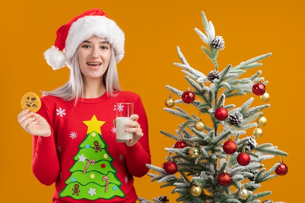 Young blonde woman in christmas sweater and santa hat holding glass of milk and cookie with smile on face