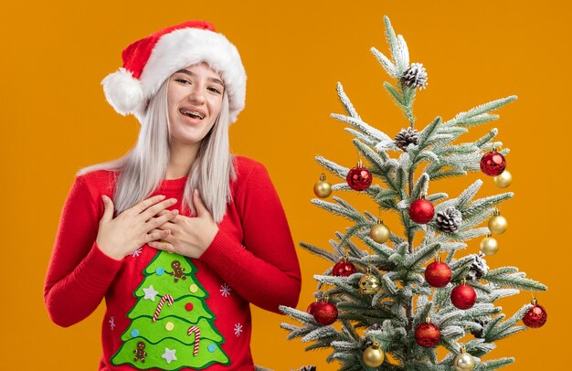 Молодая блондинка в рождественском свитере и шляпе санта-клауса, взявшись за руки на груди, чувствует благодарность, счастье и позитив