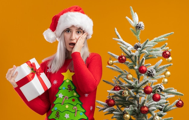 Young blonde woman in christmas  sweater and santa hat holding a present amazed  