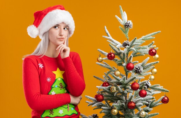 クリスマスセーターとサンタの帽子をかぶって困惑している若いブロンドの女性