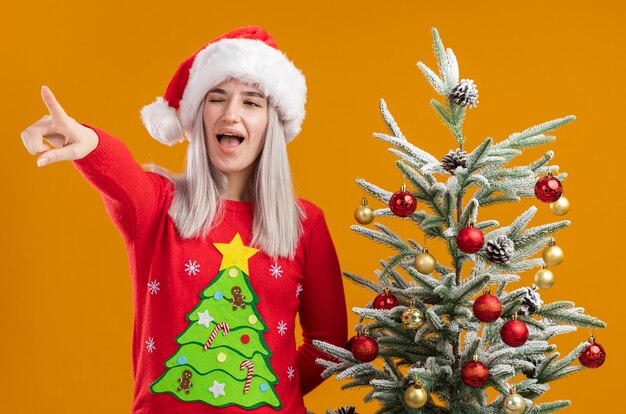 молодая блондинка в рождественском свитере и новогодней шапке смотрит в сторону, удивленно указывая указательным пальцем на что-то стоящее рядом с елкой на оранжевом фоне