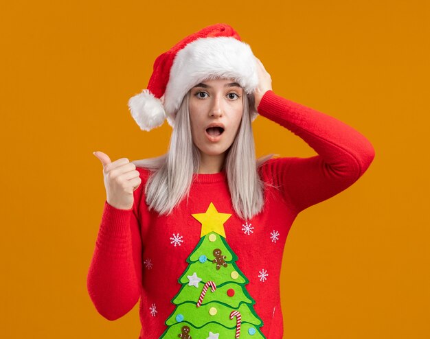 크리스마스 스웨터와 산타 모자에 젊은 금발의 여자는 오렌지 배경 위에 엄지 손가락으로 다시 가리키는 놀란 카메라를보고