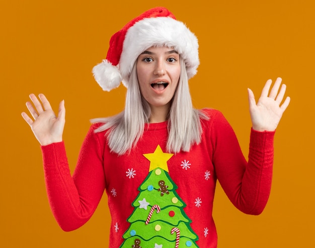 オレンジ色の背景の上に立っている手を上げて幸せで興奮してカメラを見てクリスマスセーターとサンタ帽子の若いブロンドの女性