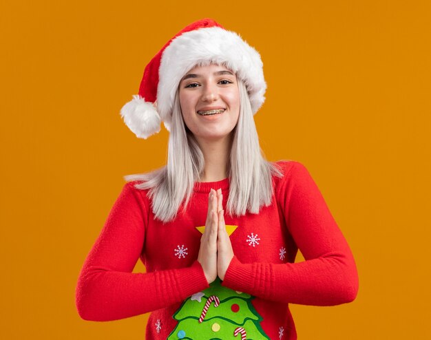 Young blonde woman in christmas sweater and santa hat looking at camera holding palms together like namaste gesture happy and positive standing over orange background