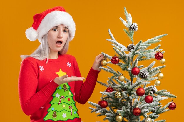 Young blonde woman in christmas  sweater and santa hat  standing next to a christmas tree confused  presenting it with arm of hand being over orange  background