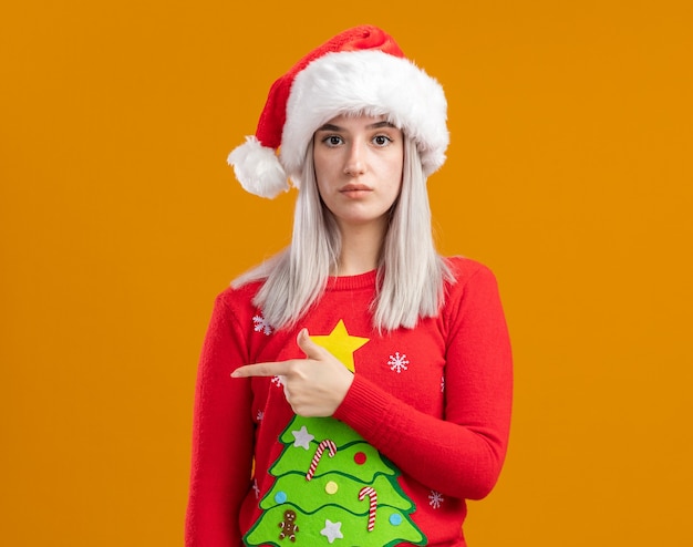 Young blonde woman in christmas  sweater and santa hat   with serious face pointing with index finger to the side  standing over orange  wall