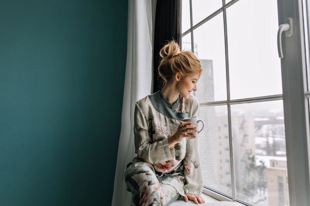 無料写真 お茶、コーヒーを飲んで、大きな窓から家でおはよう、幸せな若いブロンドの女性。花とシルクのパジャマを着ています。ターコイズ壁