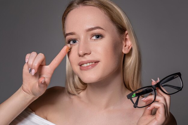 Young blonde woman holding contact lens on finger in front of her face and holding in her other hand a black glasses on gray background. Eyesight and eye care concept. Concept of choice