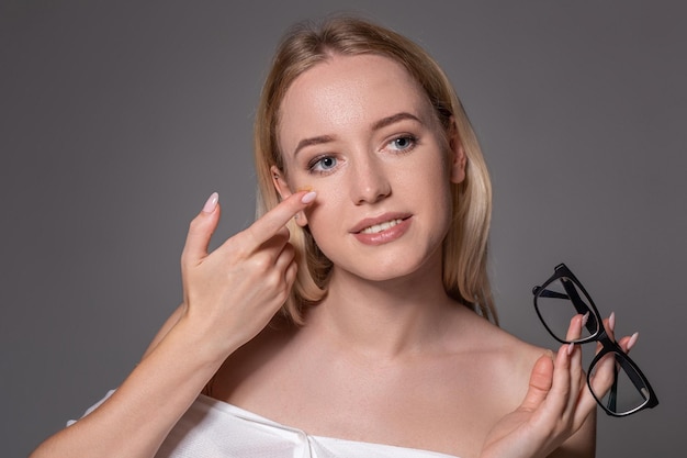 Free photo young blonde woman holding contact lens on finger in front of her face and holding in her other hand a black glasses on gray background. eyesight and eye care concept. concept of choice