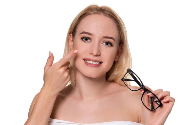 Young blonde woman holding contact lens on finger in front of her face and holding in her other hand a black glasses on white background. Eyesight and eye care concept. Concept of choice