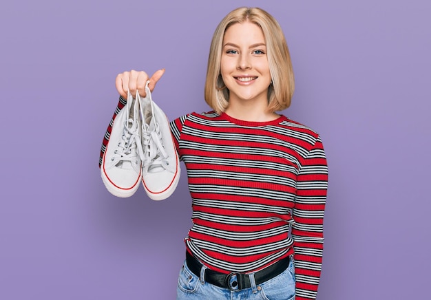 Free photo young blonde woman holding white casual shoes looking positive and happy standing and smiling with a confident smile showing teeth