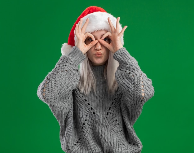 무료 사진 겨울 스웨터와 산타 모자에 젊은 금발의 여자는 녹색 배경 위에 쌍안경 제스처를 행복하고 긍정적 인 서 손가락을 통해 찾고