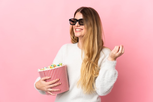 Young blonde woman over isolated wall with 3d glasses and holding a big bucket of popcorns while looking side