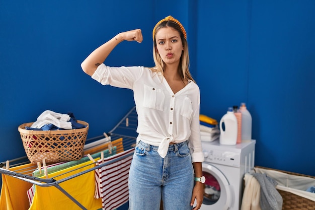 Free photo young blonde woman at laundry room strong person showing arm muscle confident and proud of power