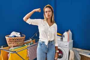 Free photo young blonde woman at laundry room strong person showing arm muscle confident and proud of power