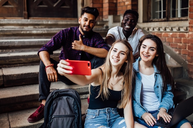 Young blonde woman making selfie with her friends students, while they are sitting on stairs.