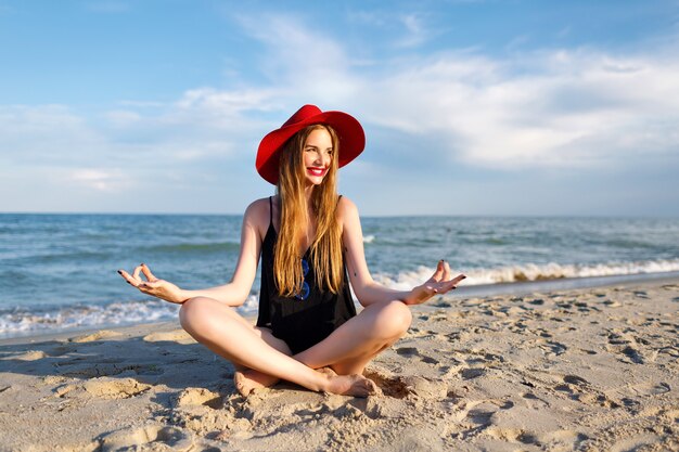 Young blonde woman meditate near ocean, bean vacation, sunshine, wearing red hat and balk top, healthy lifestyle, yoga mood. Sitting on sand and enjoy vacation.