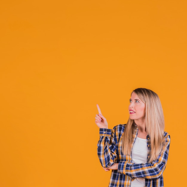 Young blonde woman pointing her finger upward looking up against an orange backdrop