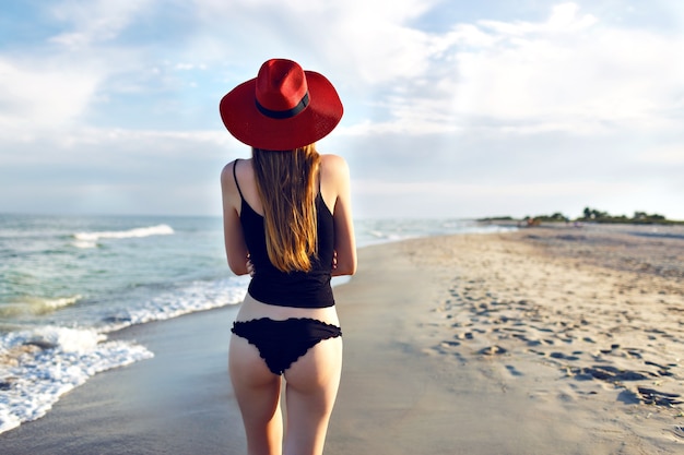 Young blonde woman posing back and walking alone near ocean, lonely beach, slim bode, wearing black bikini and elegant red hat, amazing nature view, travel on the beach.