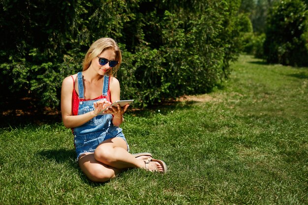 Young blonde woman sitting in park and using tablet