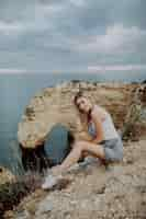 Free photo young blonde woman sitting on the rocks top by the ocean.