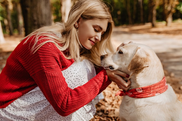 彼女の犬に微笑んでいる若いブロンドの女性。公園でペットと良い瞬間を共有するかわいい女の子。