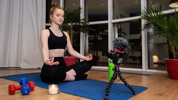 Young blonde woman in sportswear is meditating on a yoga mat looking into recording