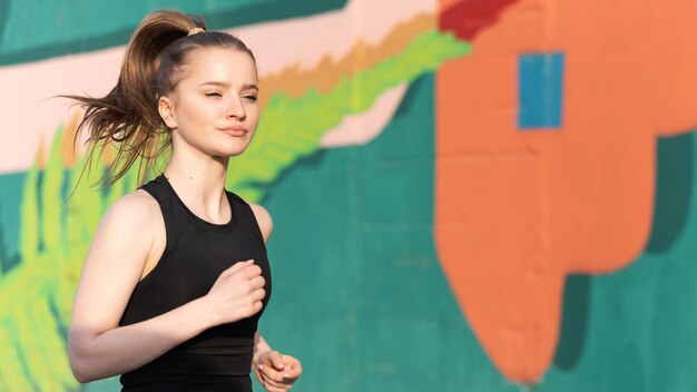 Young blonde woman in sportswear running on the road at outdoors training, multicolored wall on the background