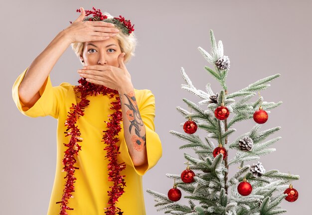 молодая блондинка в рождественском венке и гирлянде из мишуры на шее стоит возле украшенной елки и смотрит, держа руки на лбу и во рту, изолированной на белой стене