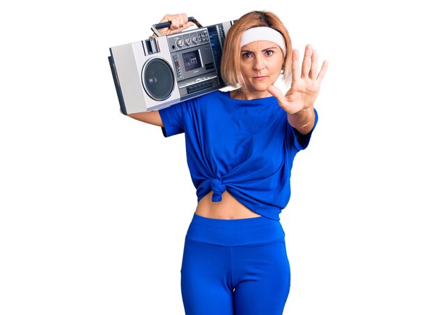 Young blonde woman wearing sportswear holding boombox, listening to music with open hand doing stop sign with serious and confident expression, defense gesture