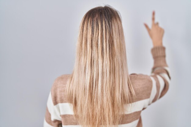 Young blonde woman wearing turtleneck sweater over isolated background posing backwards pointing ahead with finger hand