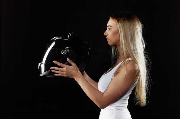young blonde woman wearing white tank top holding black motor helmet. Attractive sporty girl posing isolated with protective equipment. Extreme sports and transportation