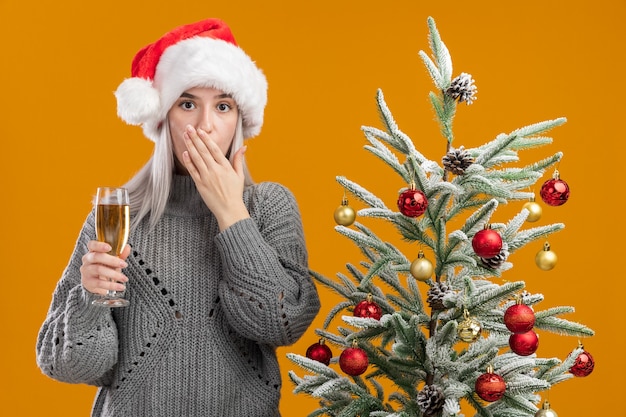 Free photo young blonde woman in winter grey  sweater and santa hat holding glass of champagne  being shocked c