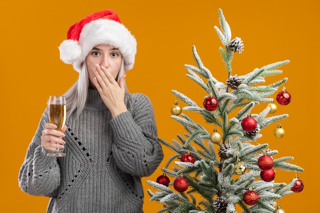 Foto gratuita giovane donna bionda in maglione grigio invernale e cappello da babbo natale con in mano un bicchiere di champagne scioccata c