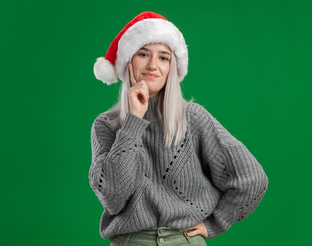 겨울 스웨터와 산타 모자에 젊은 금발의 여자 행복하고 긍정적 인 녹색 벽 위에 자신감 서 웃고