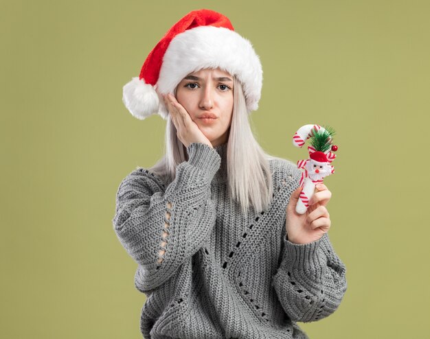 緑の壁の上に立って混乱しているクリスマスキャンディケインを保持している冬のセーターとサンタ帽子の若いブロンドの女性