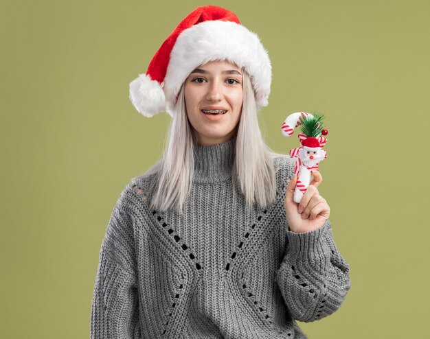 Молодая блондинка в зимнем свитере и шляпе санта-клауса держит рождественскую конфету, счастливая и взволнованная, стоя над зеленой стеной