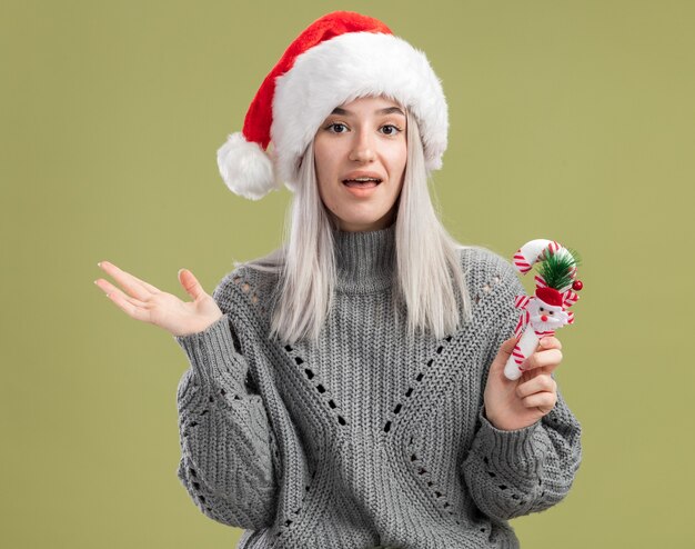 Young blonde woman in winter sweater and santa hat holding christmas candy cane  with happy face standing over green wall