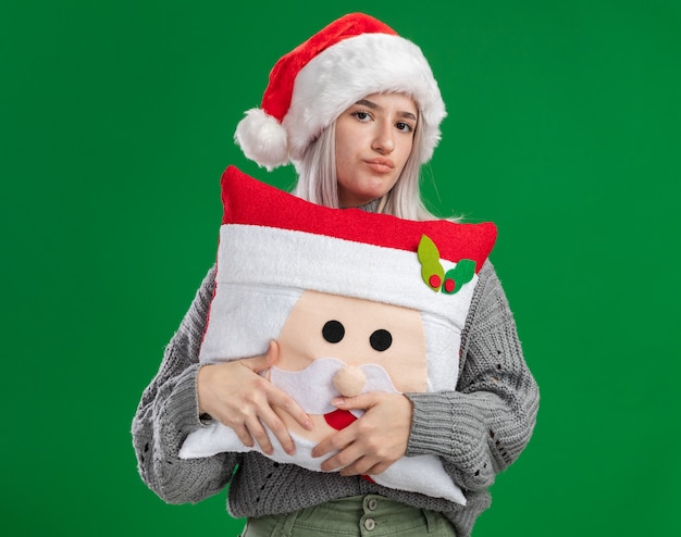 Foto gratuita giovane donna bionda in maglione invernale e cappello da babbo natale tenendo il cuscino di natale guardando la fotocamera con la fronte accigliata essendo malato in piedi su sfondo verde